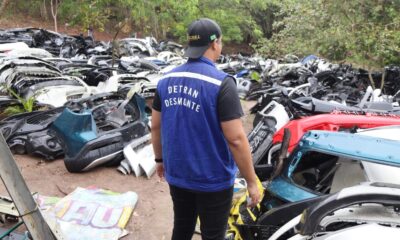 Agente de colete DETRAN DESMONTE e boné preto com bandeira do Brasil em pátio com peças automotivas diversas