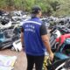 Agente de colete DETRAN DESMONTE e boné preto com bandeira do Brasil em pátio com peças automotivas diversas