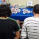 Dois homens de costas, com pilhas de pacotes, garrafas e dois rádios em mesa azul