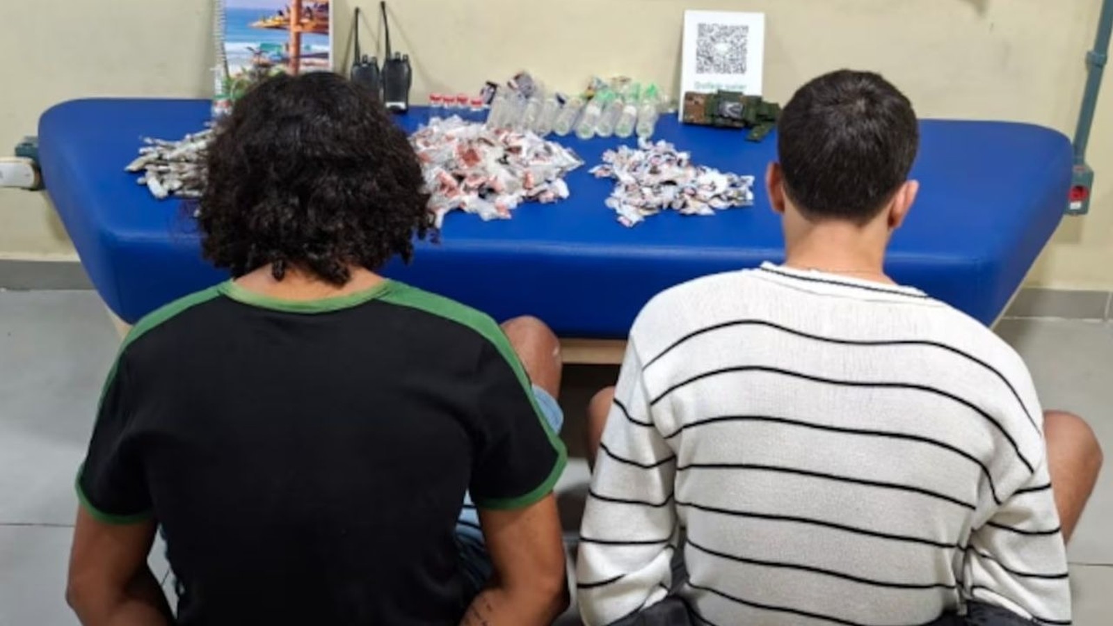 Dois homens de costas, com pilhas de pacotes, garrafas e dois rádios em mesa azul