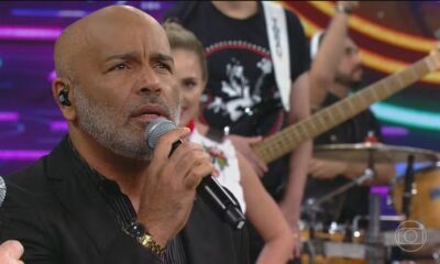 Homem careca com barba grisalha canta em microfone palco com banda desfocada atrás