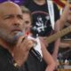Homem careca com barba grisalha canta em microfone palco com banda desfocada atrás