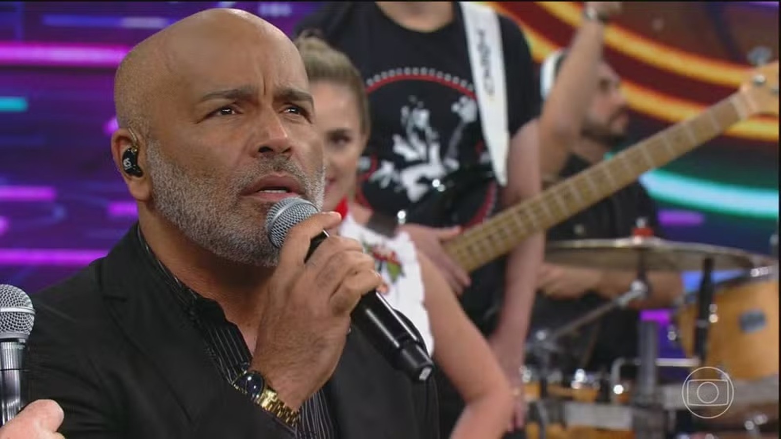 Homem careca com barba grisalha canta em microfone palco com banda desfocada atrás