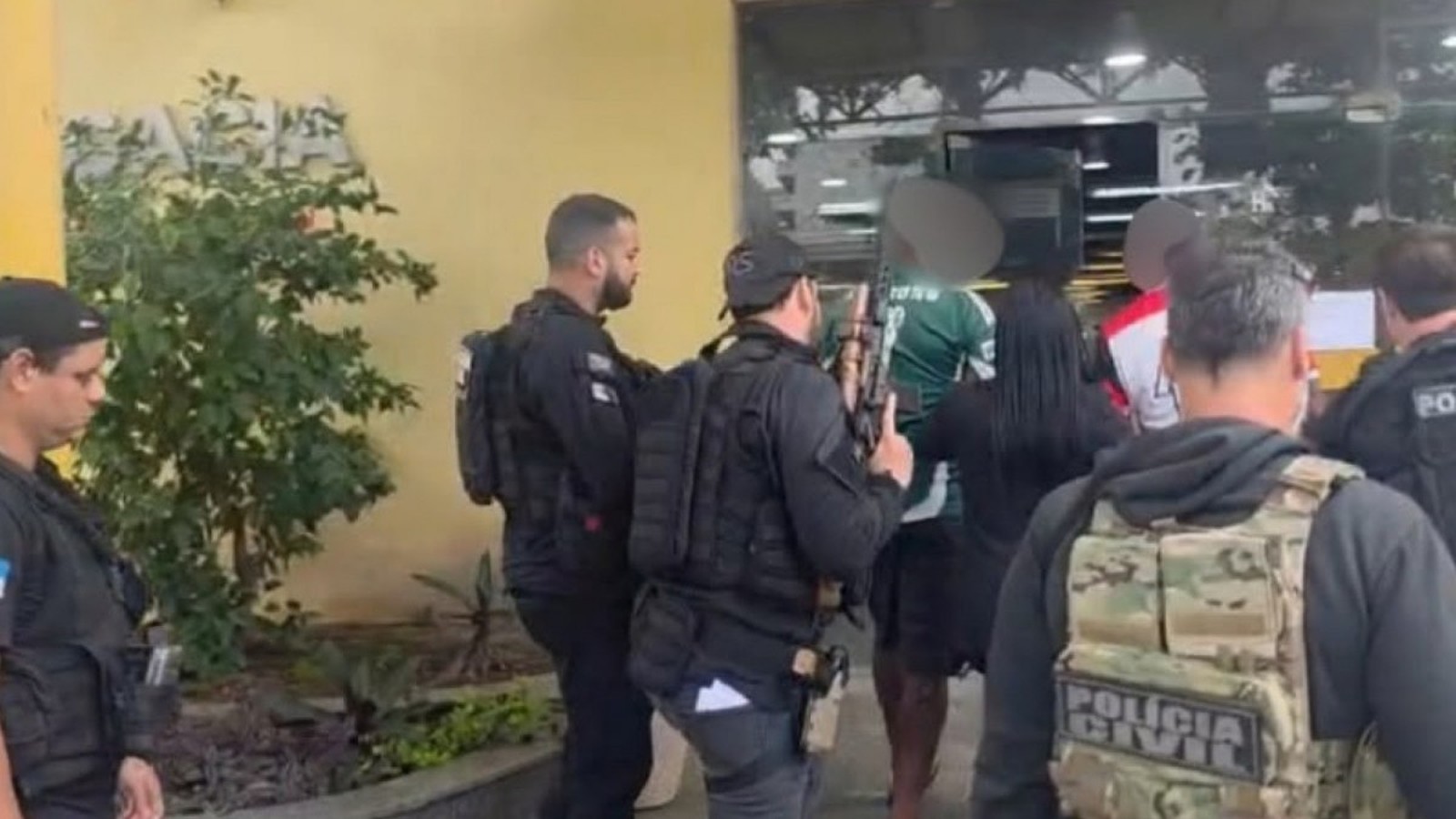 Policiais civis armados com coletes "POLÍCIA CIVIL" escoltam homens em frente a um prédio