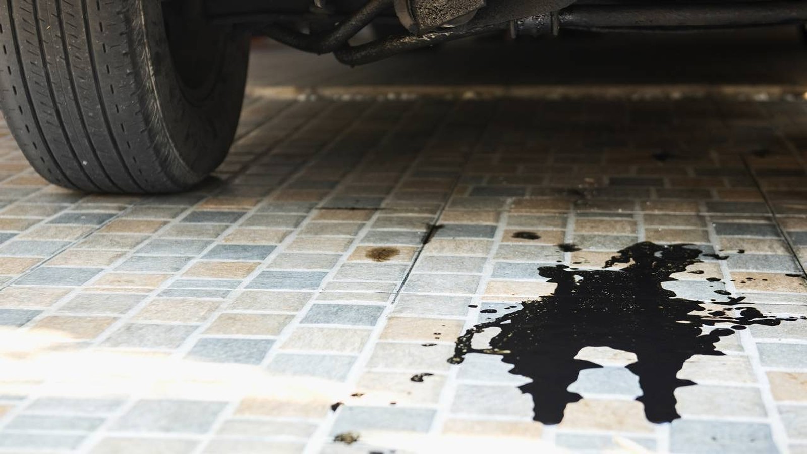 Mancha de óleo escura e grande no chão de azulejos claros, sob a roda e chassi de um carro