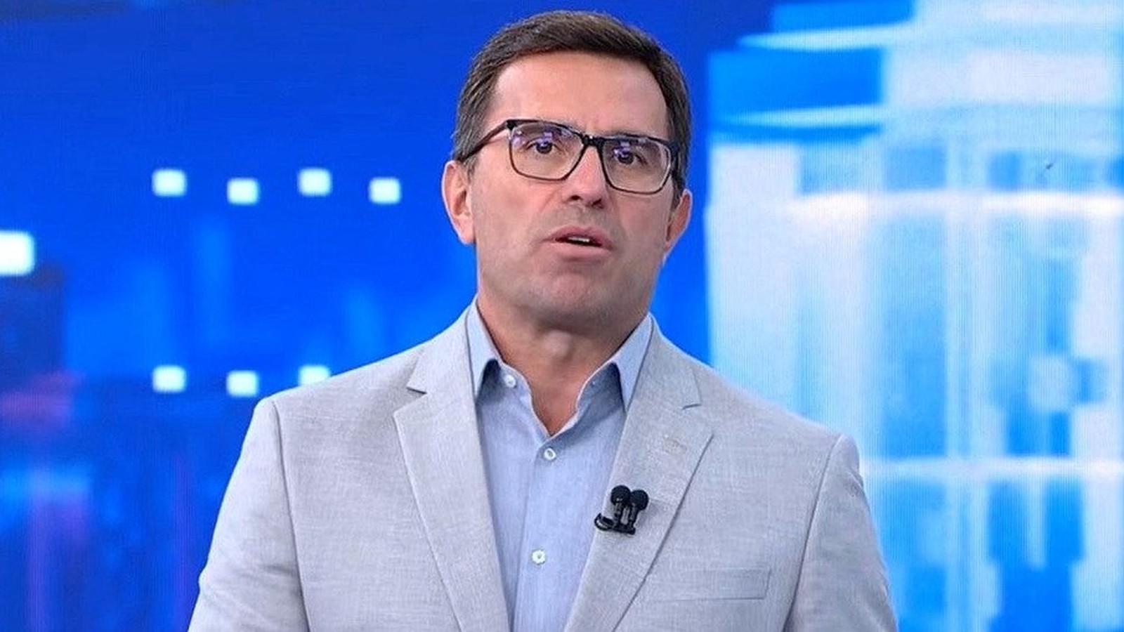 Rodrigo Bocardi, de óculos, camisa azul e paletó cinza, falando em estúdio de TV