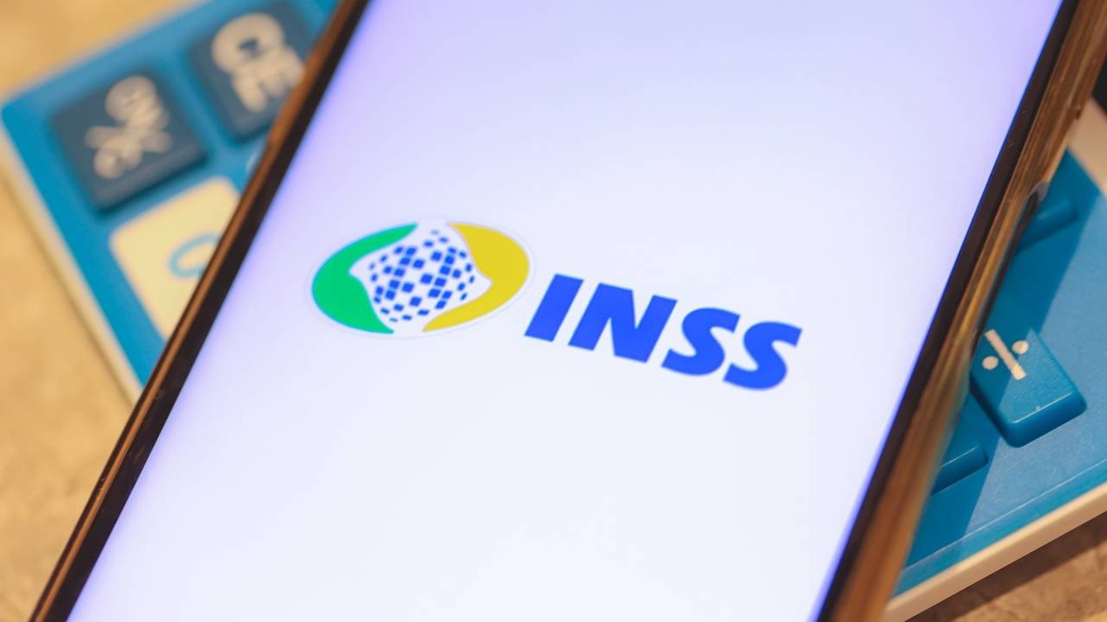 Tela de smartphone mostra o logo e as letras 'INSS' em azul, com calculadora azul no fundo desfocado