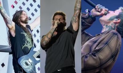 Três músicos masculinos tatuados no palco: um guitarrista e dois vocalistas cantando