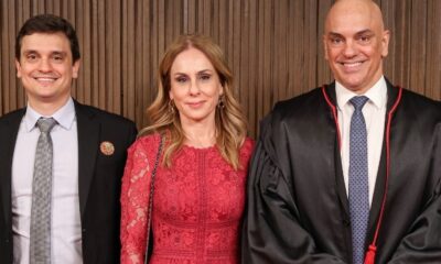 Três pessoas sorrindo: um homem de terno, uma mulher de vestido vermelho e um homem de toga.