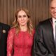 Três pessoas sorrindo: um homem de terno, uma mulher de vestido vermelho e um homem de toga.