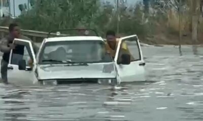 Dois homens em meio à água alta abrem as portas de um carro branco submerso em enchente