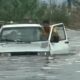 Dois homens em meio à água alta abrem as portas de um carro branco submerso em enchente