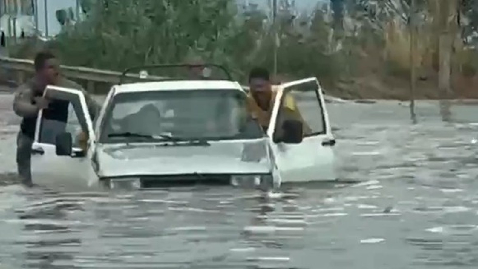 Dois homens em meio à água alta abrem as portas de um carro branco submerso em enchente