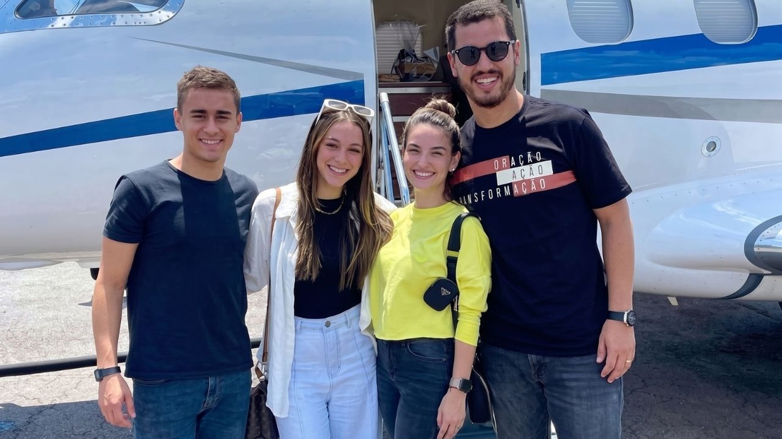 Quatro jovens sorrindo em frente a um jato particular branco e azul