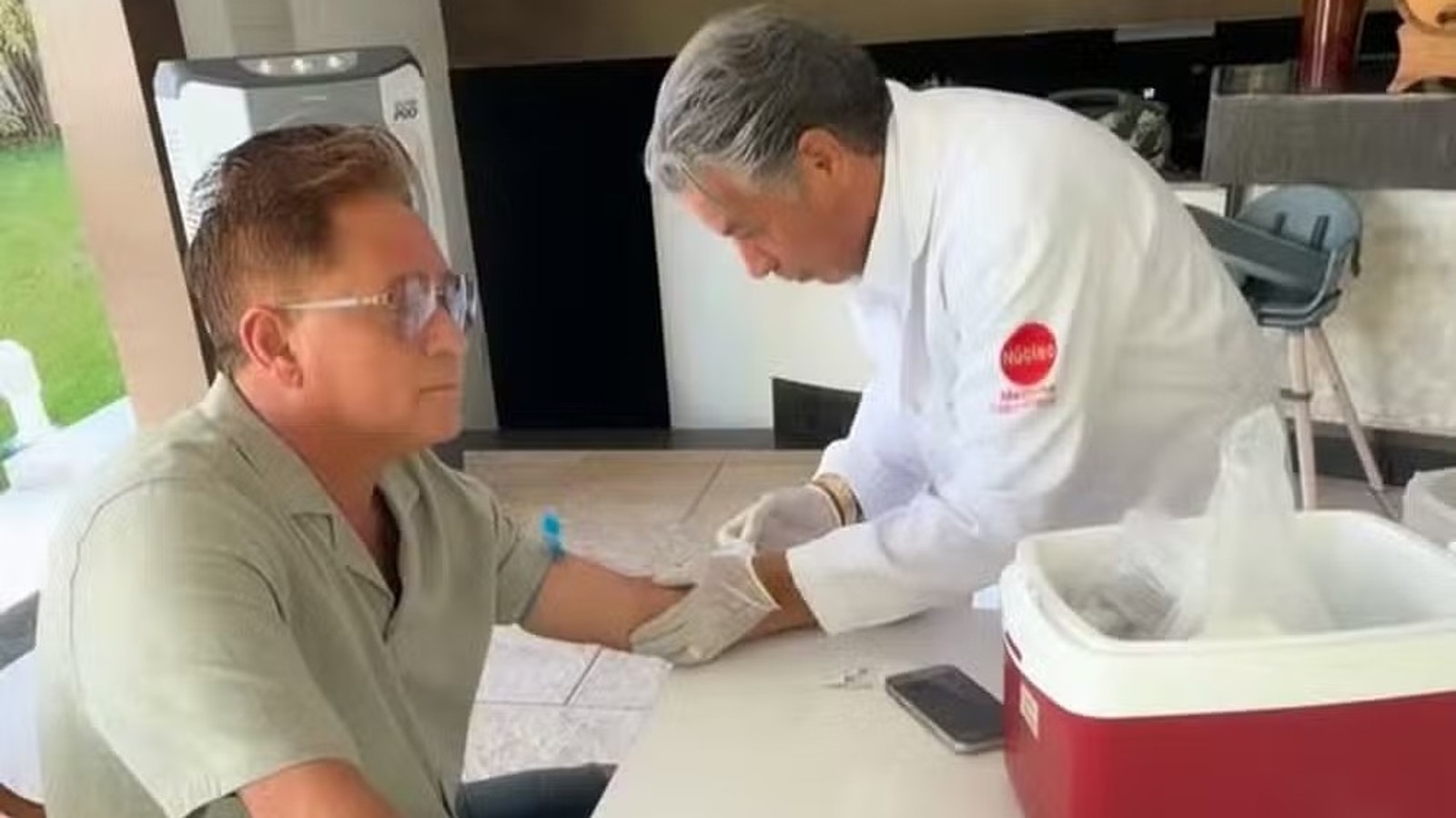Leonardo, de óculos, tem sangue coletado do braço por profissional de jaleco branco