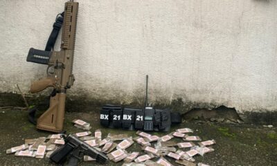 Fuzil, pistola, rádio, cinto tático com 'BX 21', e drogas em pacotes no chão