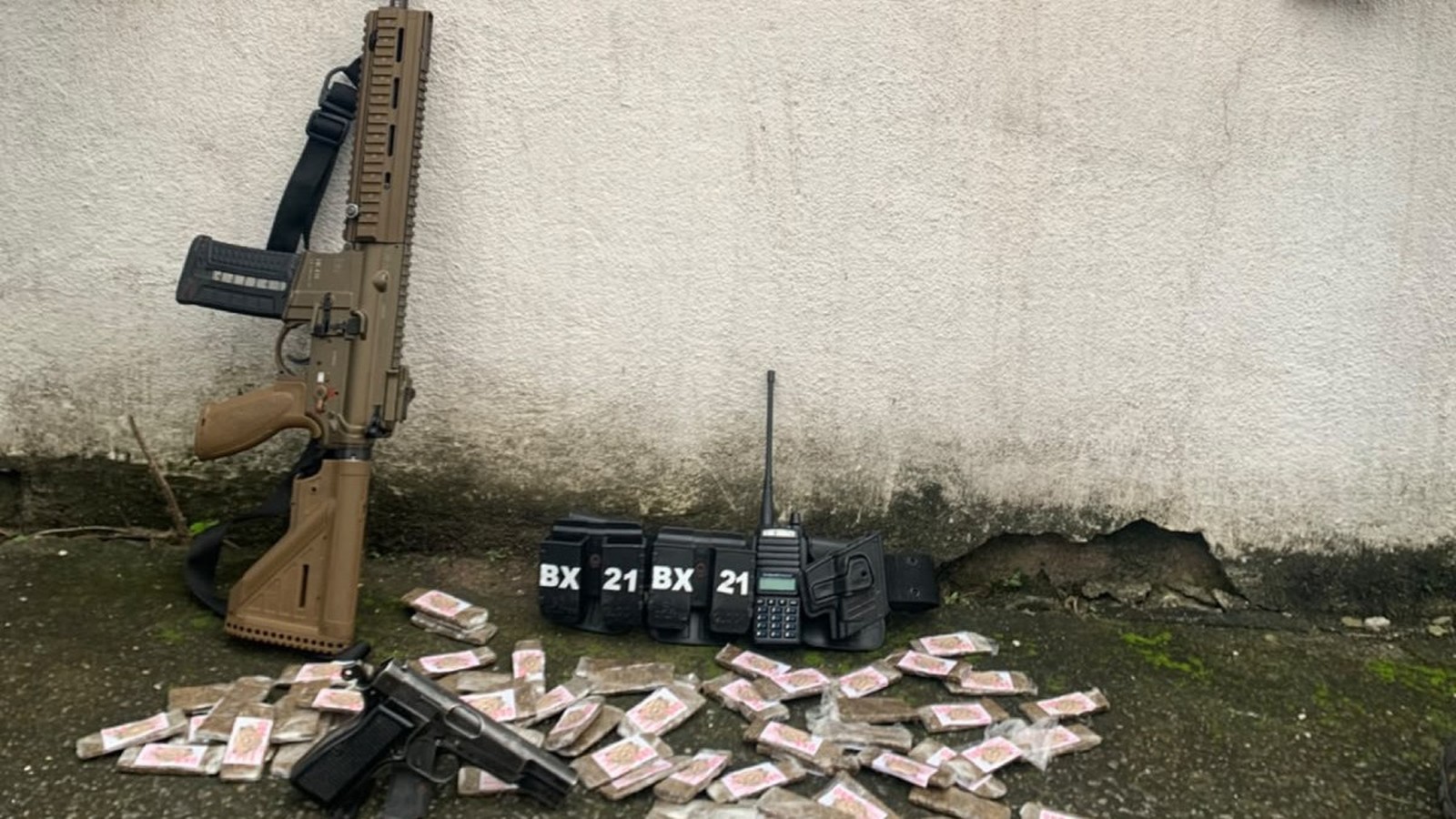 Fuzil, pistola, rádio, cinto tático com 'BX 21', e drogas em pacotes no chão