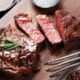 Bife grelhado suculento com alecrim em tábua de madeira, ao lado de sal grosso e pimenta-do-reino