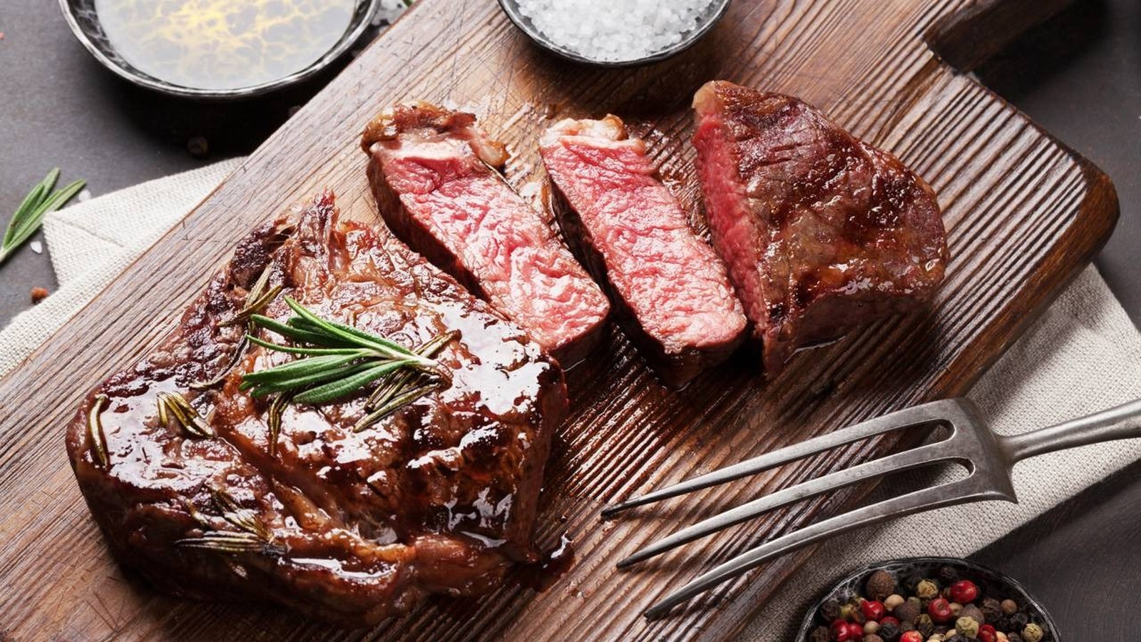 Bife grelhado suculento com alecrim em tábua de madeira, ao lado de sal grosso e pimenta-do-reino