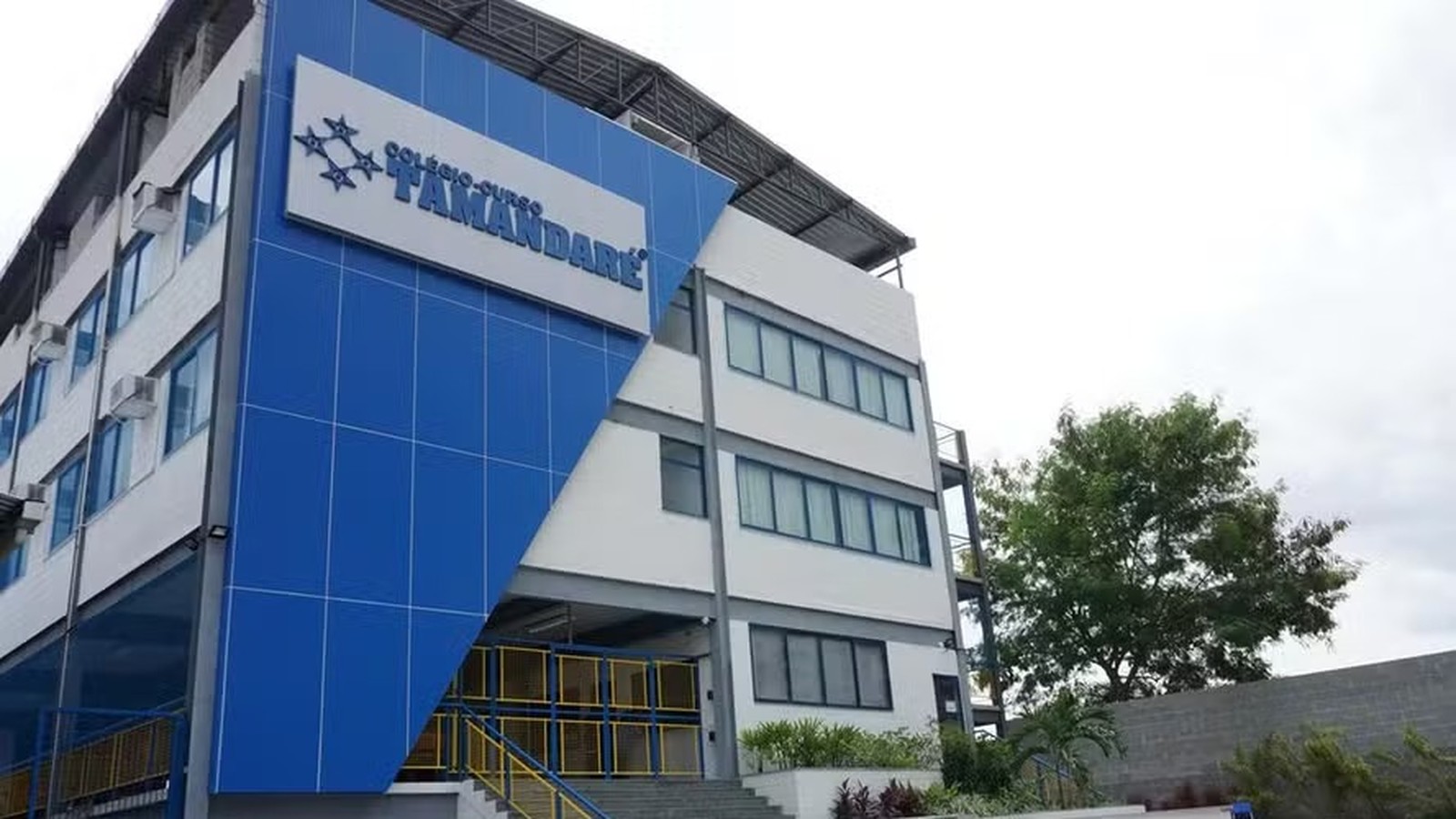 Edifício moderno com fachada azul e branca, placa "COLÉGIO-CURSO TAMANDARÉ", janelas e entrada com escadas