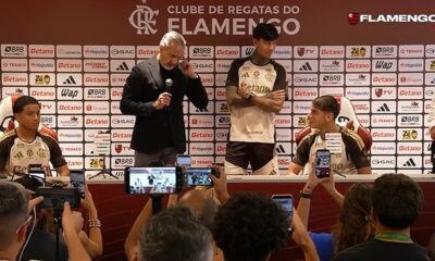 Diretor e dois jovens jogadores em coletiva de imprensa com fundo de patrocinadores e pessoas filmando com celulares