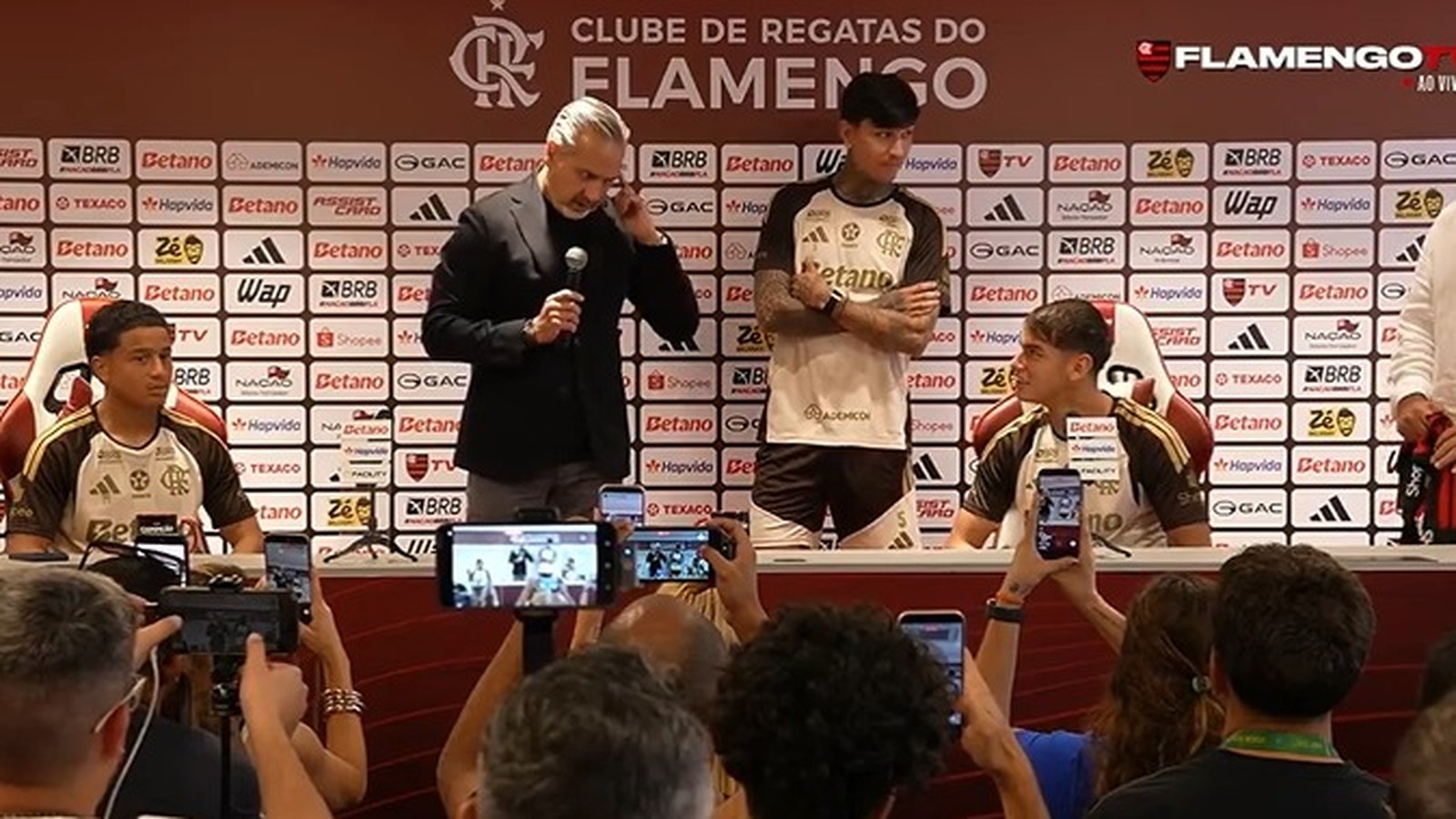 Diretor e dois jovens jogadores em coletiva de imprensa com fundo de patrocinadores e pessoas filmando com celulares