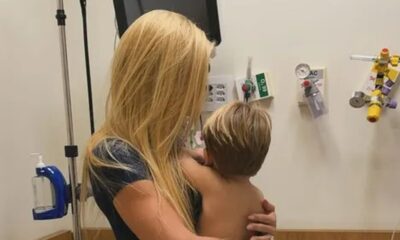 Mulher loira, de costas, segura uma criança loira de costas em quarto de hospital com equipamentos.
