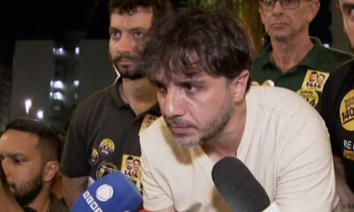 Homem barbudo com microfone em evento noturno, cercado por apoiadores com broches políticos.