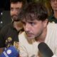Homem barbudo com microfone em evento noturno, cercado por apoiadores com broches políticos.