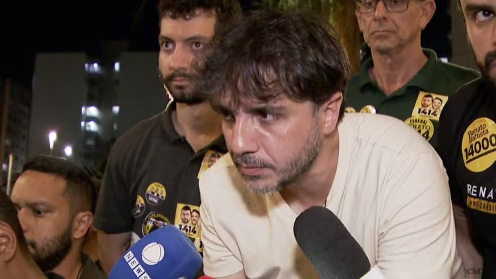 Homem barbudo com microfone em evento noturno, cercado por apoiadores com broches políticos.