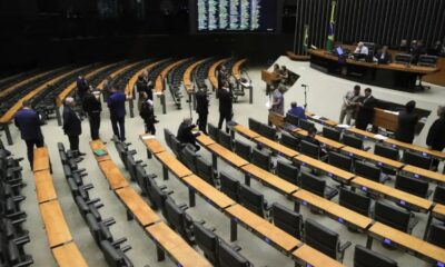 Plenário de assembleia legislativa com cadeiras e mesas, homens em ternos conversando, e tela grande ao fundo