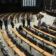 Plenário de assembleia legislativa com cadeiras e mesas, homens em ternos conversando, e tela grande ao fundo
