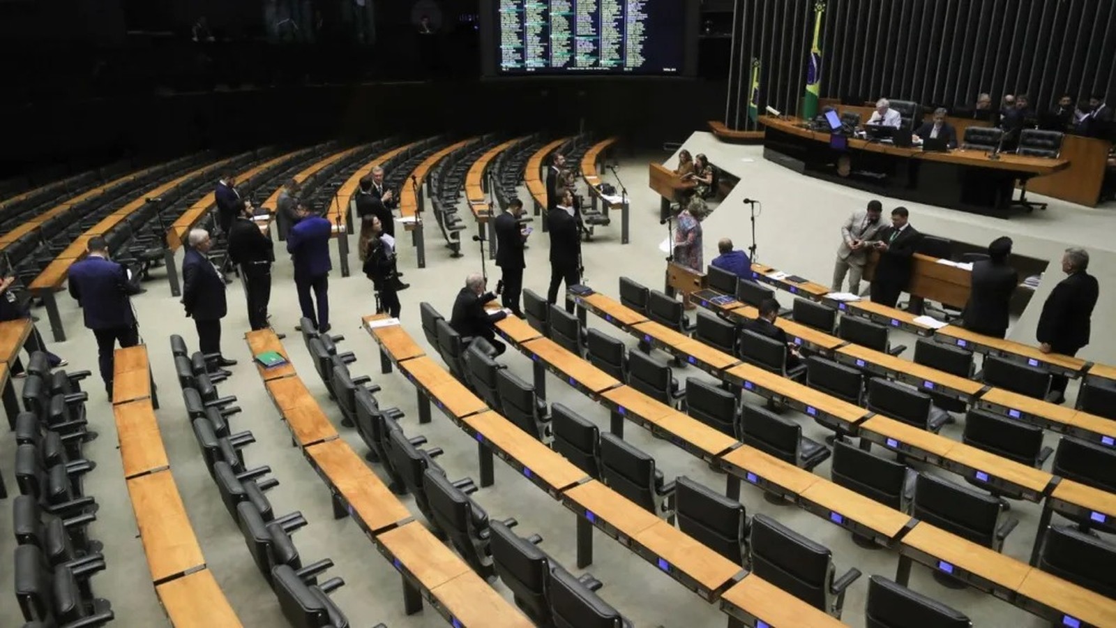 Plenário de assembleia legislativa com cadeiras e mesas, homens em ternos conversando, e tela grande ao fundo