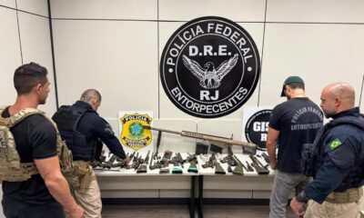 Policiais examinam fuzil calibre .50 e diversas armas apreendidas sobre uma mesa