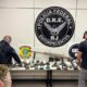 Policiais examinam fuzil calibre .50 e diversas armas apreendidas sobre uma mesa