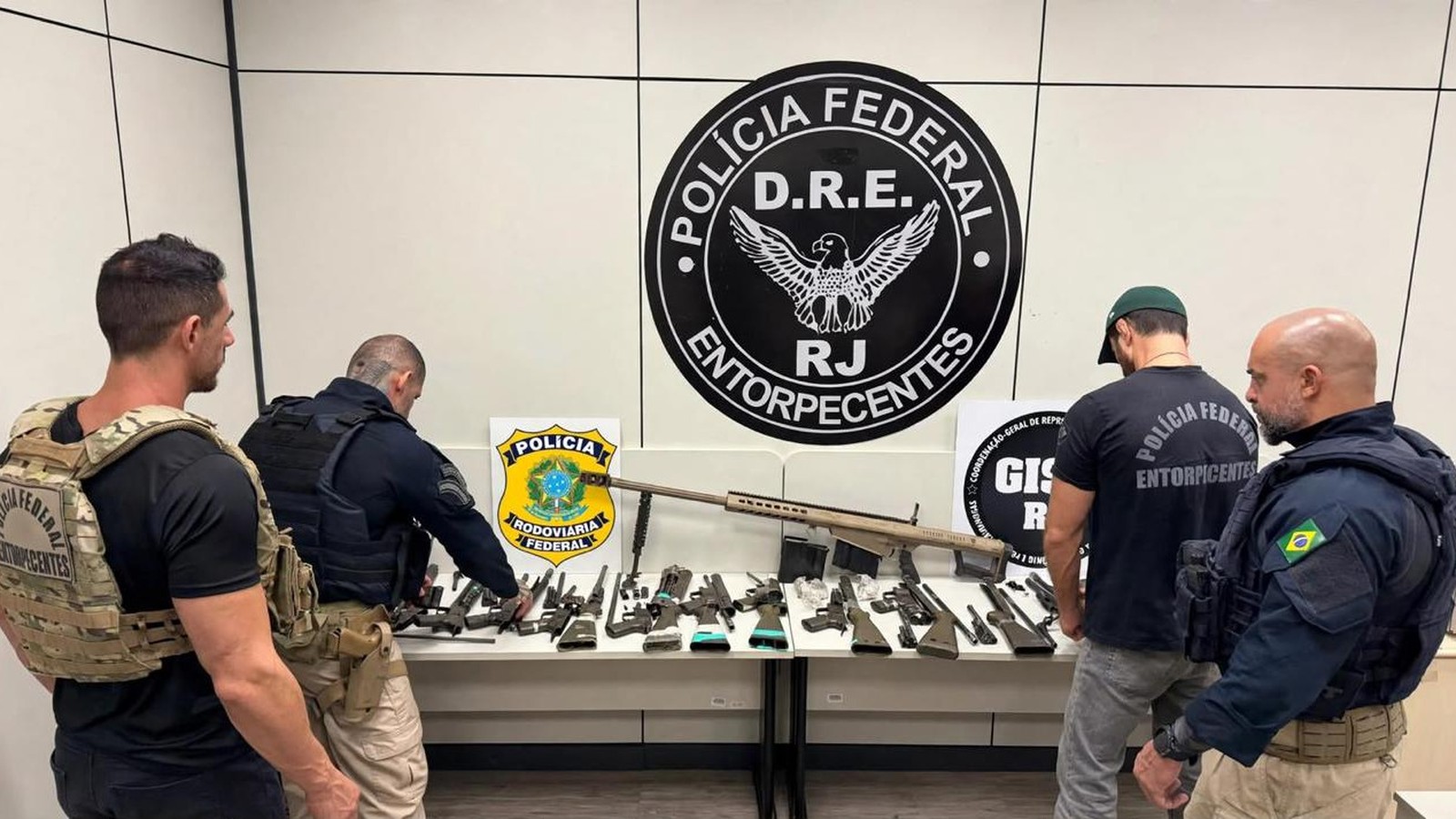 Policiais examinam fuzil calibre .50 e diversas armas apreendidas sobre uma mesa