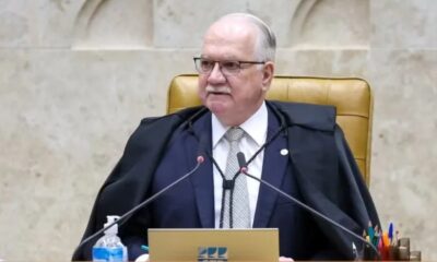 Homem de óculos, terno e toga sentado à mesa com microfones e álcool em gel
