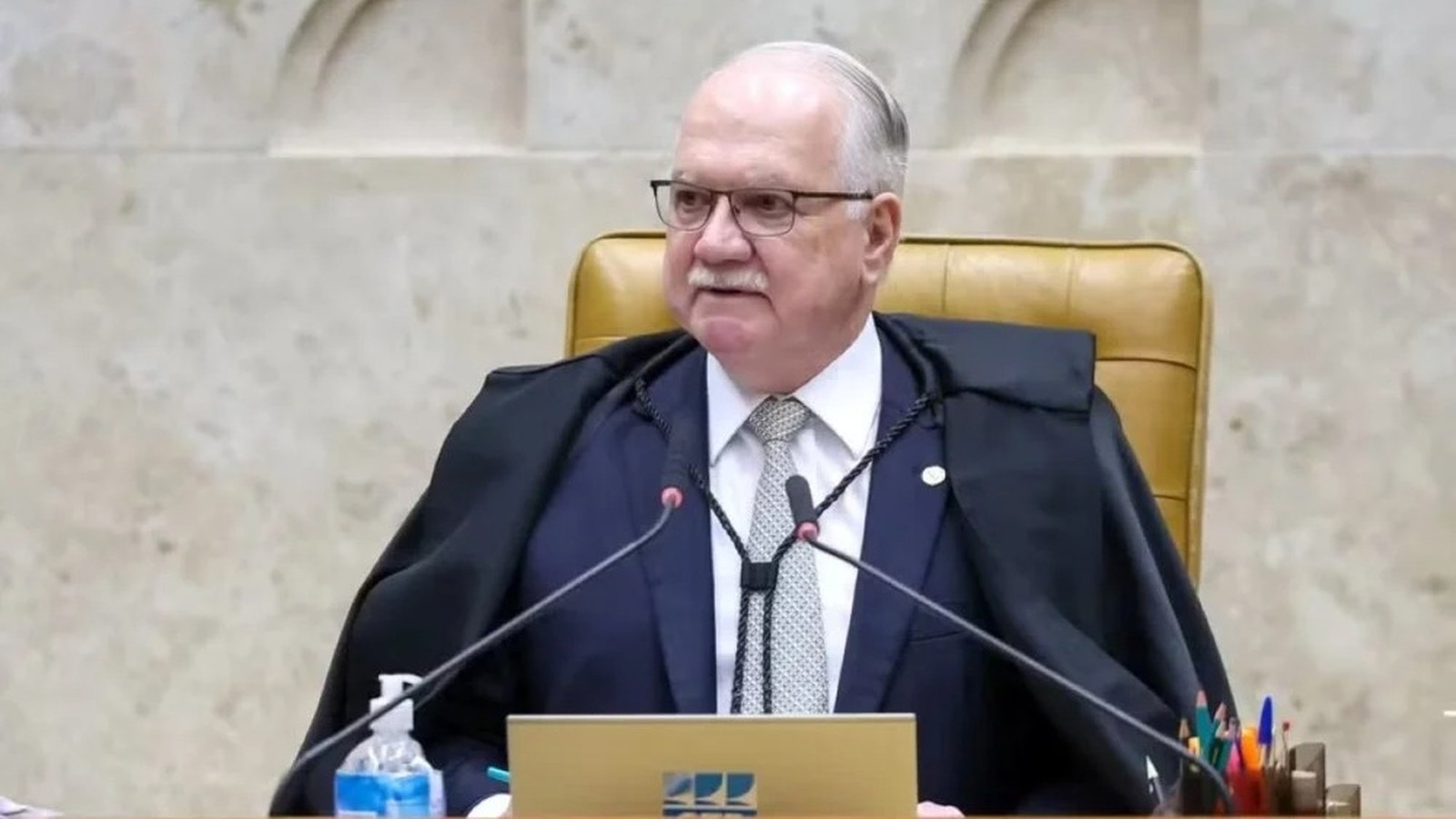 Homem de óculos, terno e toga sentado à mesa com microfones e álcool em gel