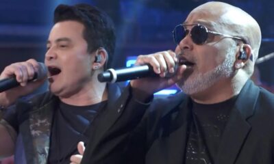 Dois homens cantando em microfones, o da direita com óculos escuros e barba