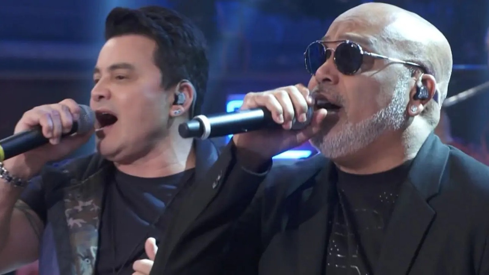 Dois homens cantando em microfones, o da direita com óculos escuros e barba