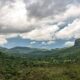 Vasto vale verde com montanhas íngremes, um paredão rochoso e céu dramático com nuvens