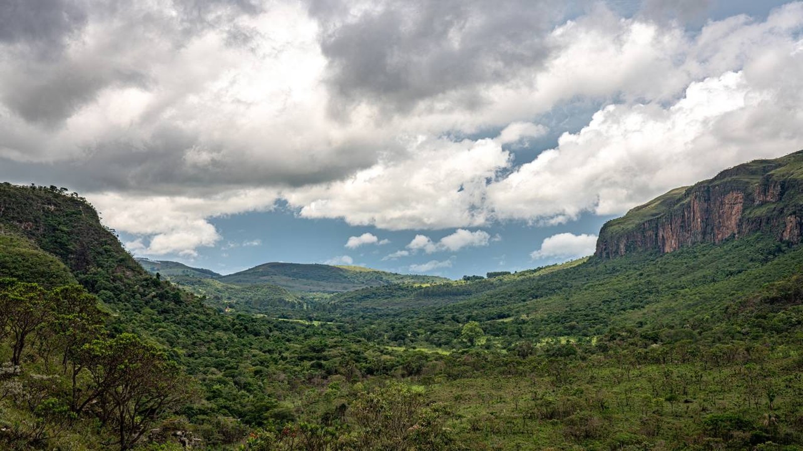 Vasto vale verde com montanhas íngremes, um paredão rochoso e céu dramático com nuvens