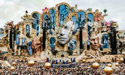 Palco de festival com arquitetura fantasiosa, rostos esculpidos, multidão e esferas douradas flutuantes