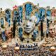Palco de festival com arquitetura fantasiosa, rostos esculpidos, multidão e esferas douradas flutuantes