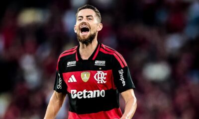 Jogador de futebol barbudo com camisa listrada preta e vermelha do Flamengo, boca aberta, expressando emoção