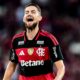 Jogador de futebol barbudo com camisa listrada preta e vermelha do Flamengo, boca aberta, expressando emoção