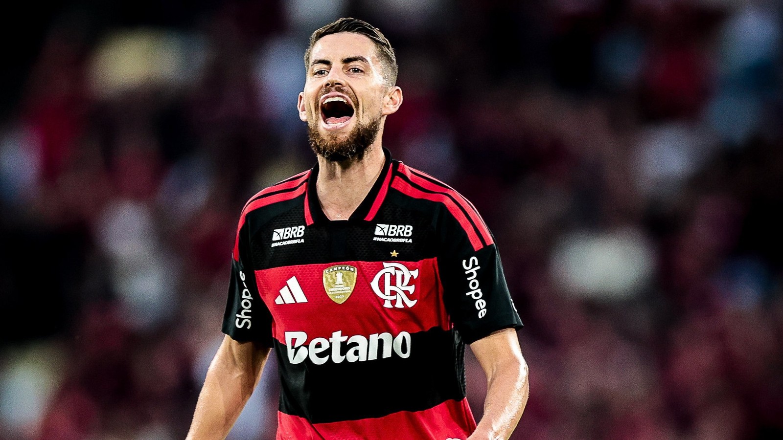 Jogador de futebol barbudo com camisa listrada preta e vermelha do Flamengo, boca aberta, expressando emoção