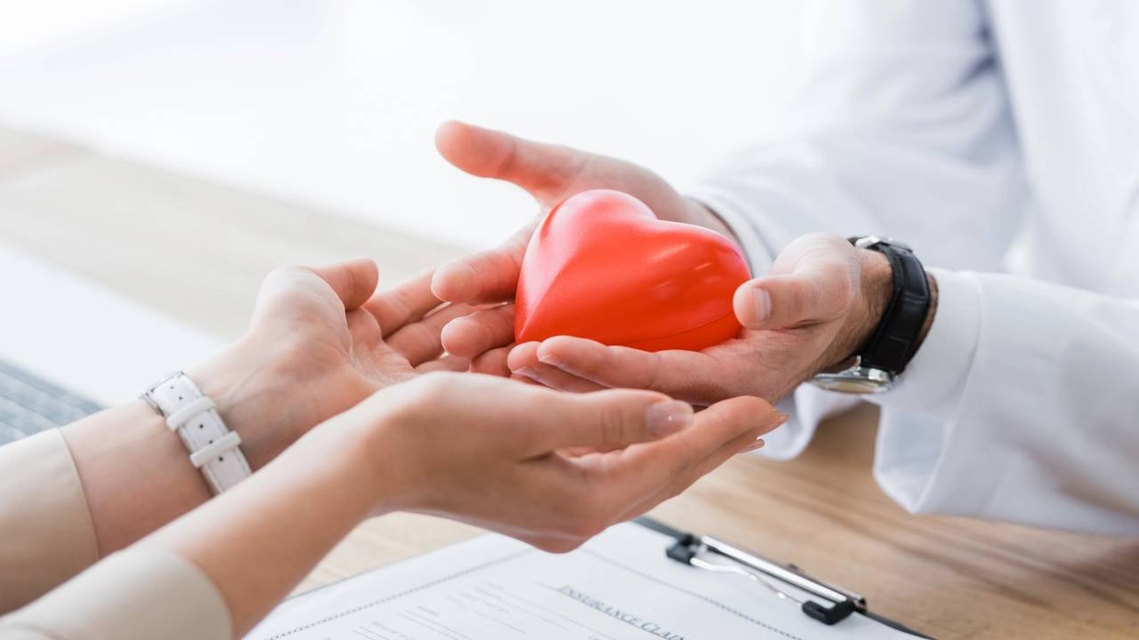 Mãos de médico e paciente seguram coração vermelho, com documento 'INSURANCE CLA' ao fundo