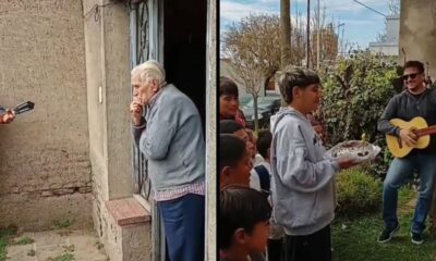 Idoso olha da porta e crianças com bolo e violonista cantam parabéns na rua