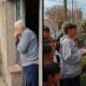 Idoso olha da porta e crianças com bolo e violonista cantam parabéns na rua